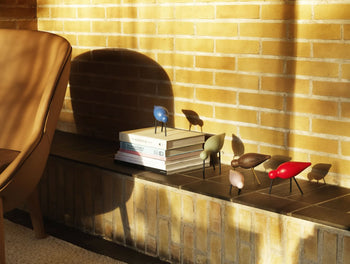The Normann Copenhagen Shorebird Small Oak by Normann Copenhagen stands on a ledge next to stacked books against a yellow brick wall, with sunlight casting deep shadows—ideal for nature-inspired decor.