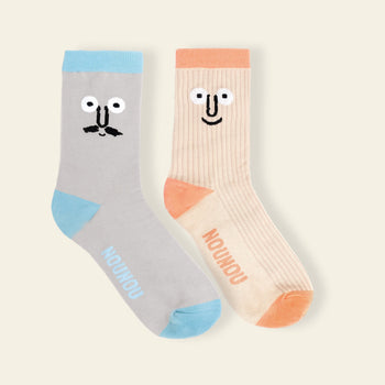 The NouNou Face Socks Set - Coral/Blue by NouNou features two playful embroidered socks with cartoon faces—one gray with a mustache and blue toe, the other beige with a smiling face, orange toe, and "NOUNOU" on the soles.