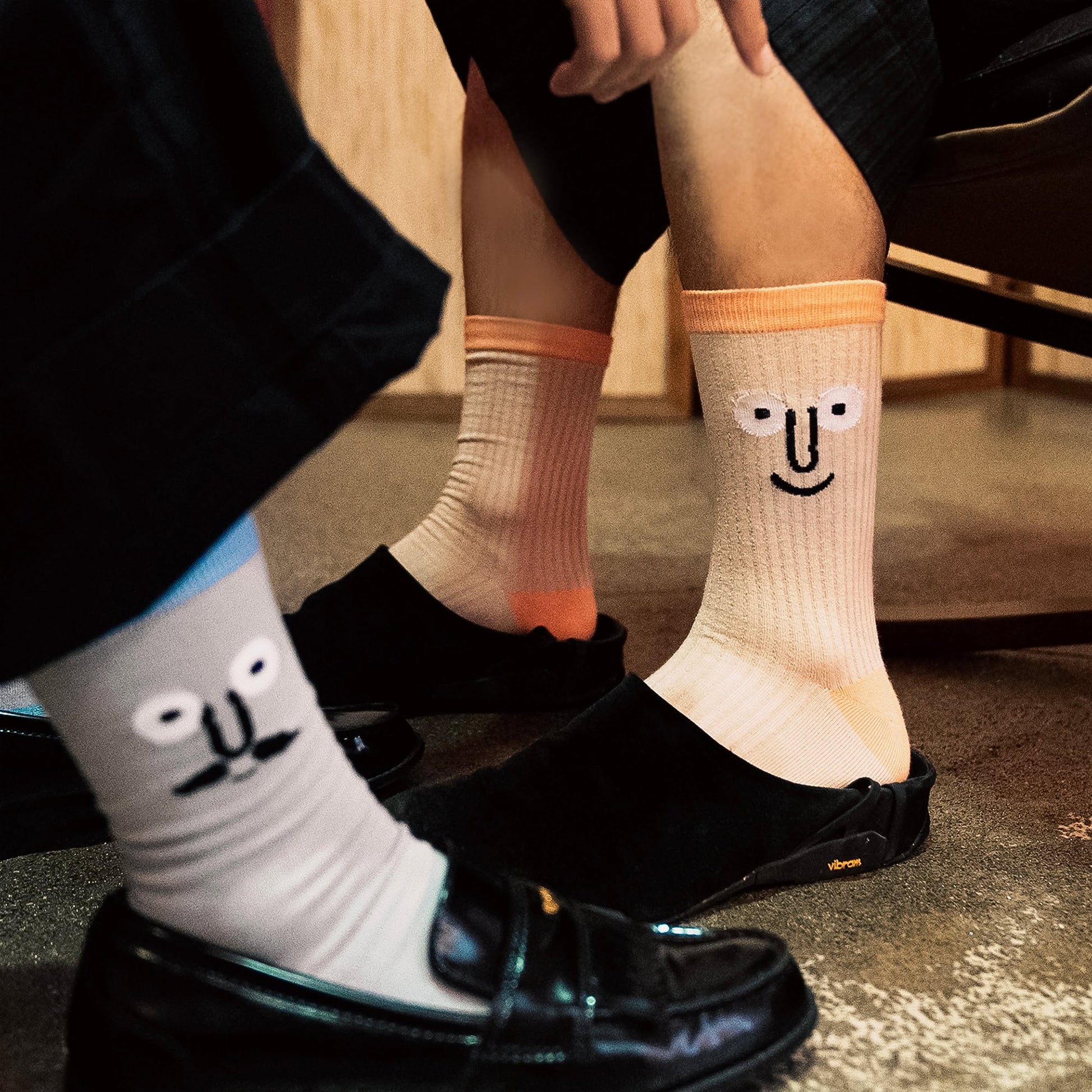 Two people sit indoors on concrete, wearing black slip-ons and the NouNou Face Socks Set - Coral/Blue by NouNou. These playful socks feature embroidered cartoon faces—a mustache with glasses and a smile—adding charm to any look.