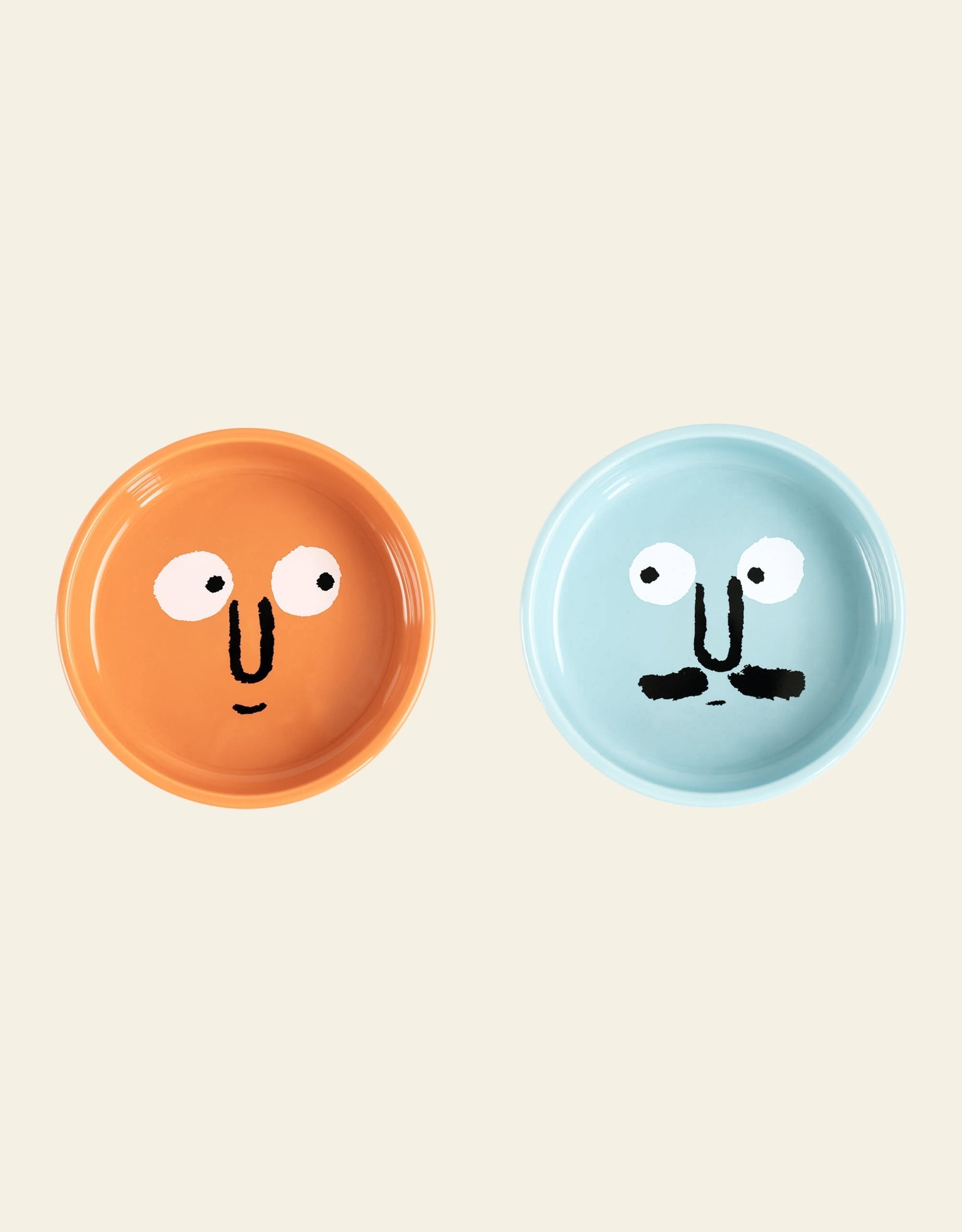 The NouNou Friends Plate Set - Mint Blue/Coral Orange by NouNou features two handmade round ceramic plates—one coral orange with a smiling face, and one mint blue with a mustache—displayed on a plain beige background.