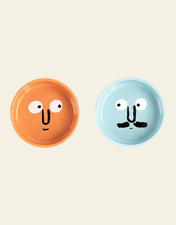 The NouNou Friends Plate Set - Mint Blue/Coral Orange by NouNou features two handmade round ceramic plates—one coral orange with a smiling face, and one mint blue with a mustache—displayed on a plain beige background.