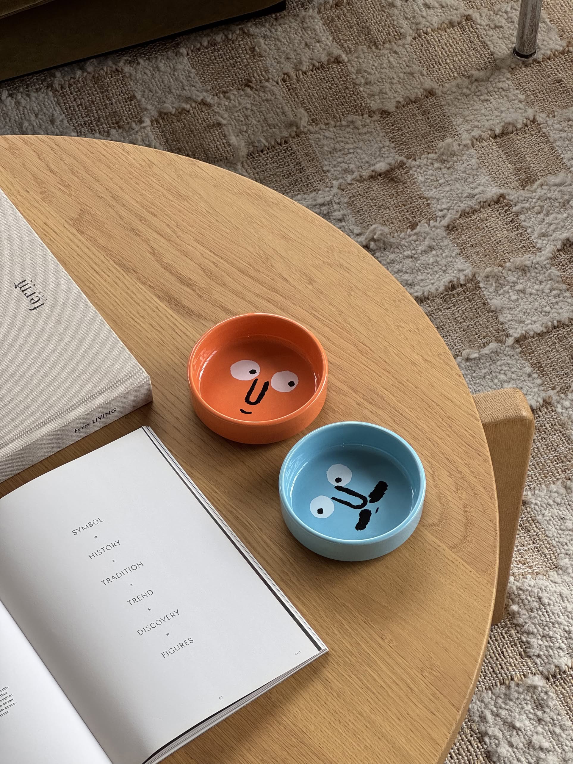 Two small handmade plates from NouNous Friends Plate Set—Mint Blue and Coral Orange, each featuring the original NouNou face—rest on a wooden coffee table beside an open and a closed book on a patterned rug.
