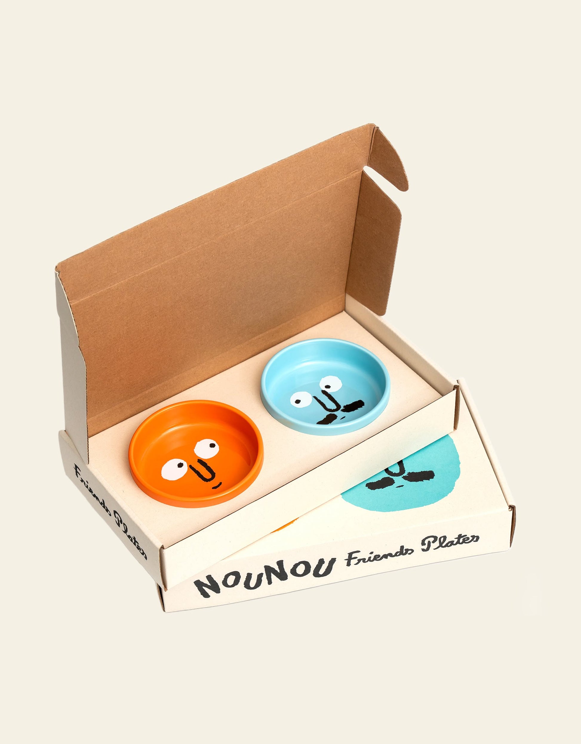 The NouNou Friends Plate Set - Mint Blue/Coral Orange by NouNou features two handmade ceramic plates, one mint blue and one coral orange, each with a whimsical smiling face, packaged in an open branded cardboard box.