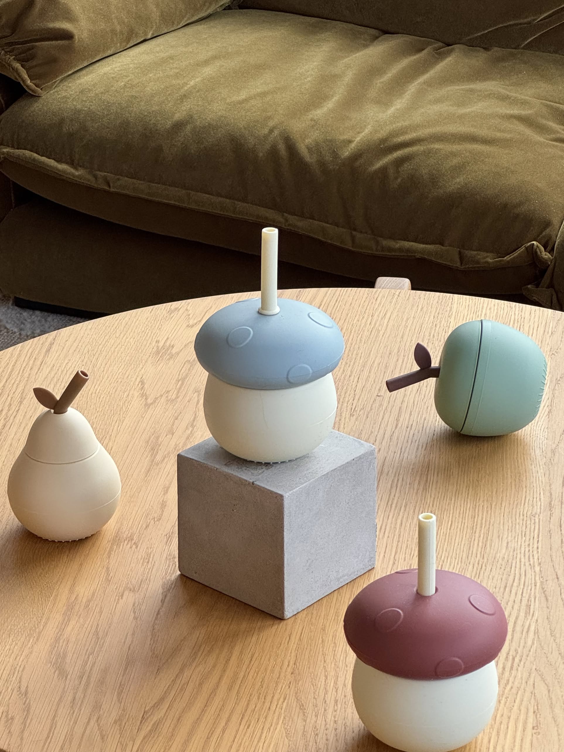 Three OYOY Living Design Mushroom Cups in pastel blue, green, and beige with straws, along with a maroon pear-shaped silicone cup, rest on a light wood table. A brown couch appears in the background.