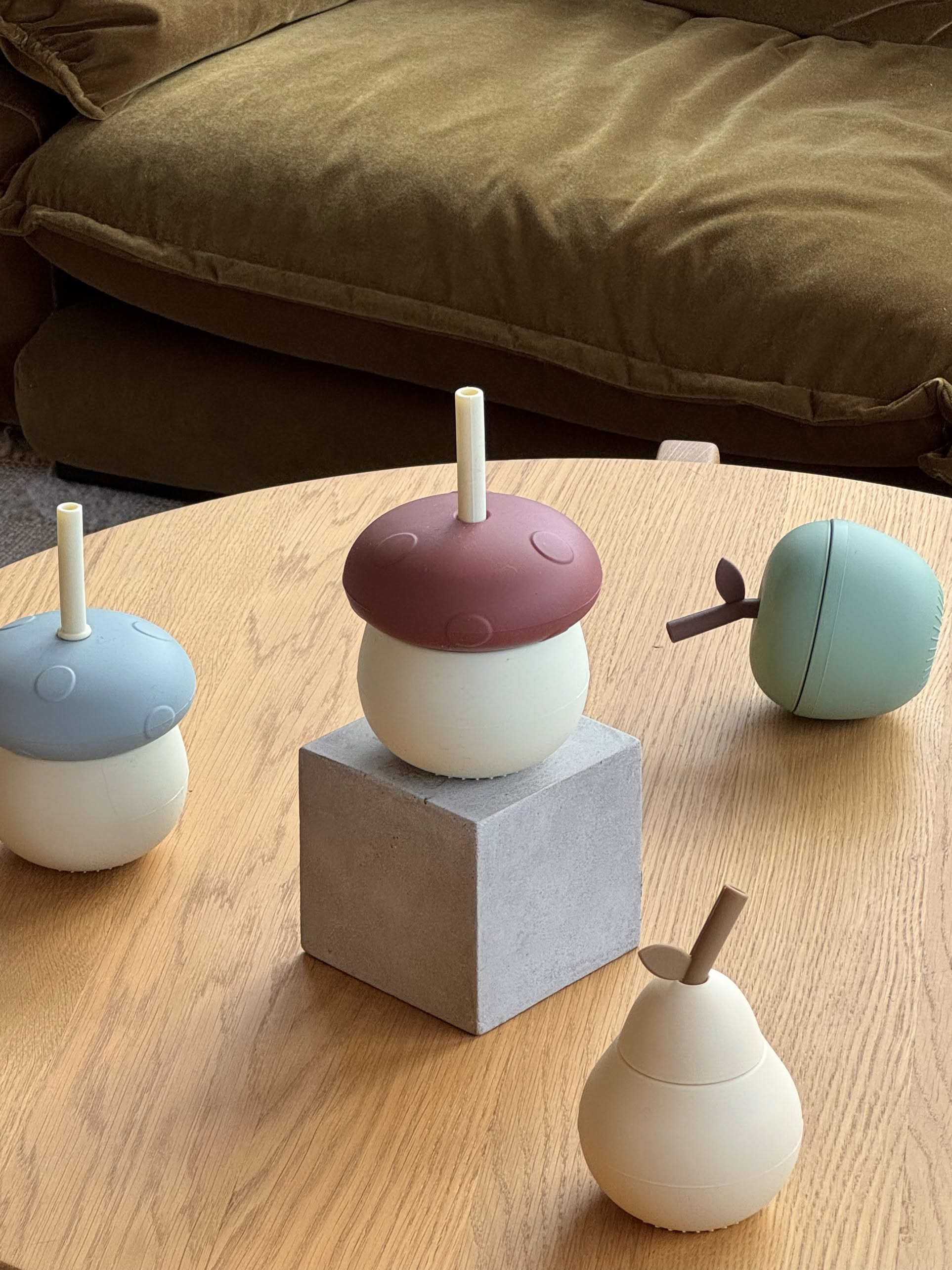 Four playful OYOY Living Design Mushroom Cups in pastel silicone, some with straws, sit on a light wooden table. One cup is elevated on a gray block, with a brown sofa in the background. Brand: OYOY Living Design.