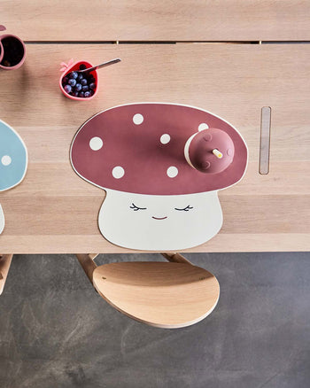 A wooden table set with a cheerful mushroom-shaped placemat, the OYOY Living Design Mushroom Cup with lid, and a pink heart-shaped dish of blueberries. A light wooden chair from OYOY Living Design is partially visible at the table.