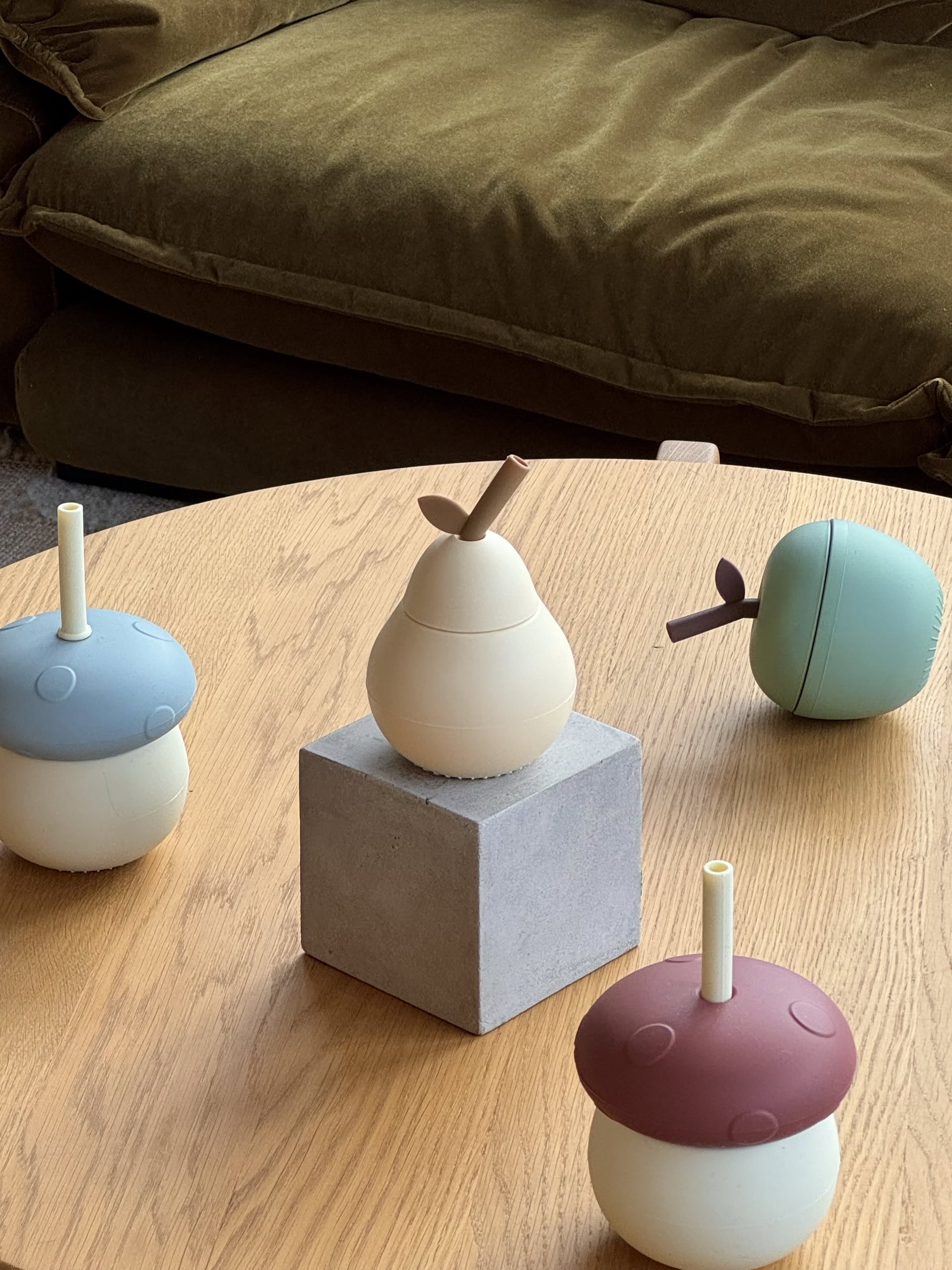 The OYOY Living Design Pear Cup - Vanilla sits on a square block, surrounded by colorful silicone mushroom cups from OYOY Living Design on a wooden table, with a brown couch in the background.