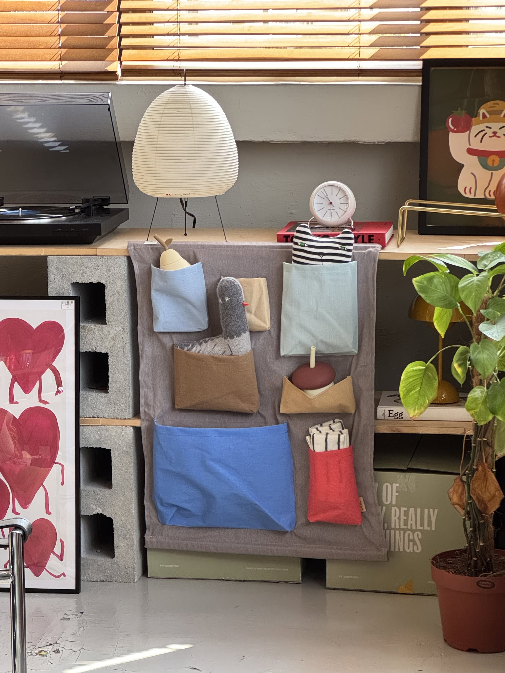 A cozy corner features the OYOY Living Design Saga Wall Storage for organizing pouches, a pigeon plush toy, and small items. Nearby sit a potted plant, turntable, artwork, clock, and sunlight filtering through window blinds.