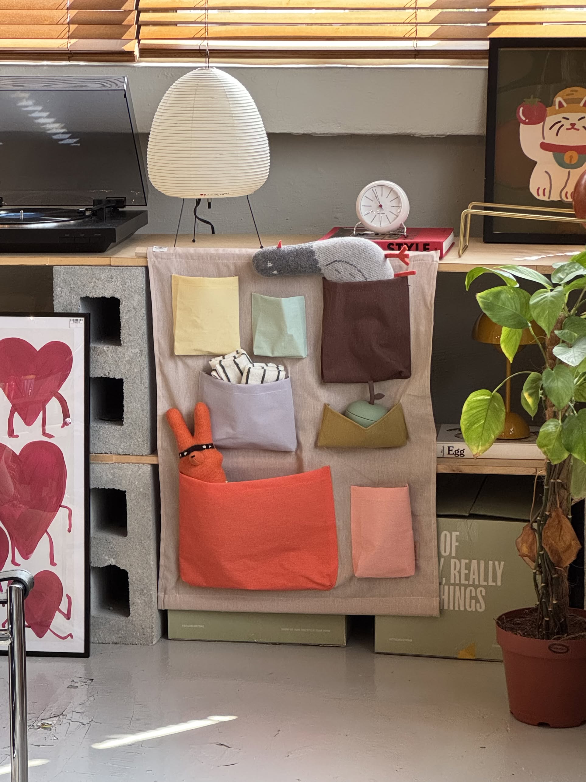 The OYOY Living Design Saga Wall Storage, made of durable fabric with colorful pockets, hangs on a shelf and holds plush toys. Beside it, sunlight streams over a potted plant, cat print, record player, and clock.
