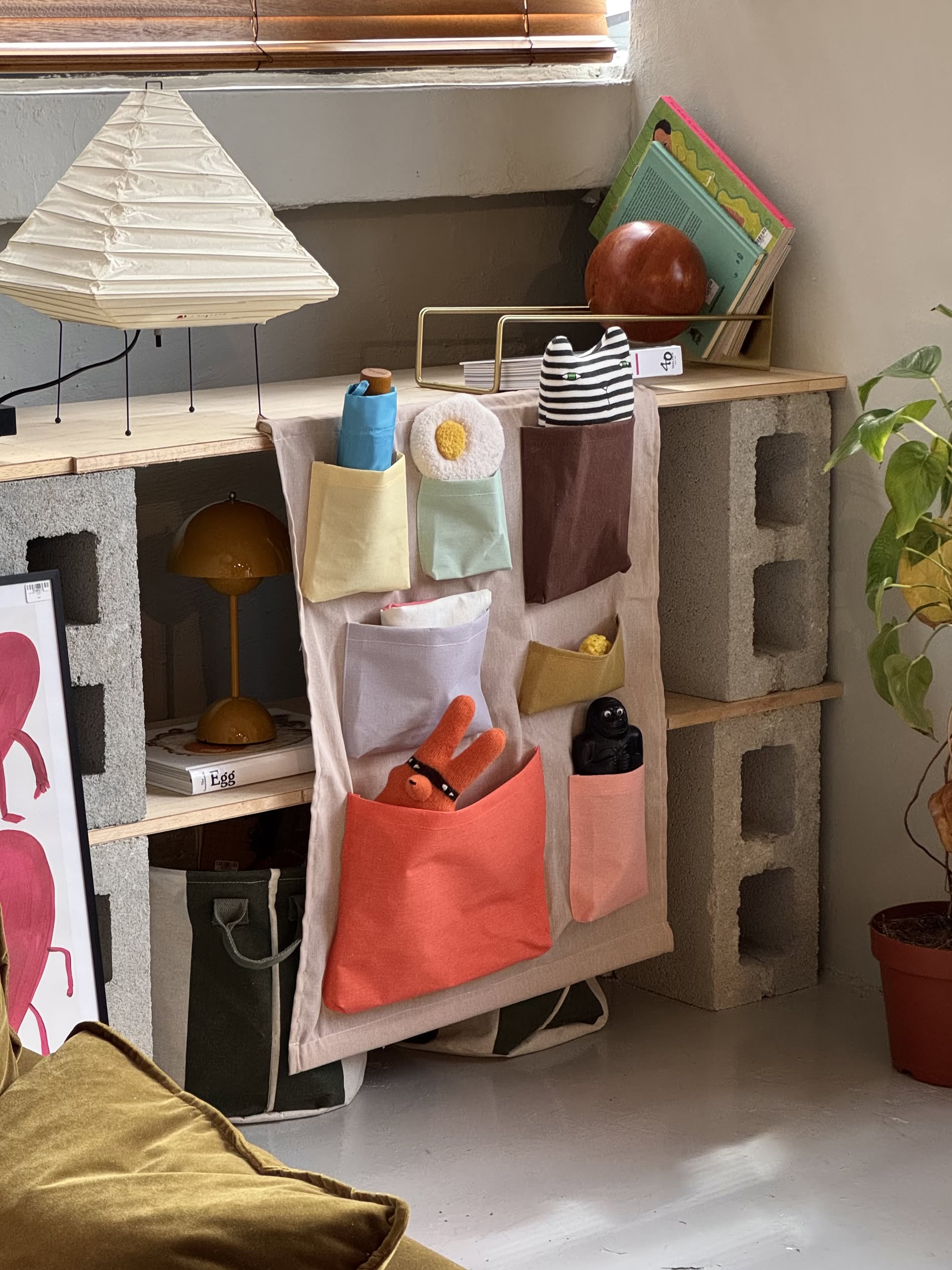 The OYOY Living Design Saga Wall Storage hangs on concrete blocks and wooden planks in a cozy, plant-filled room. Its colorful, durable fabric pockets hold plush toys, a striped stuffed animal, and other small items.