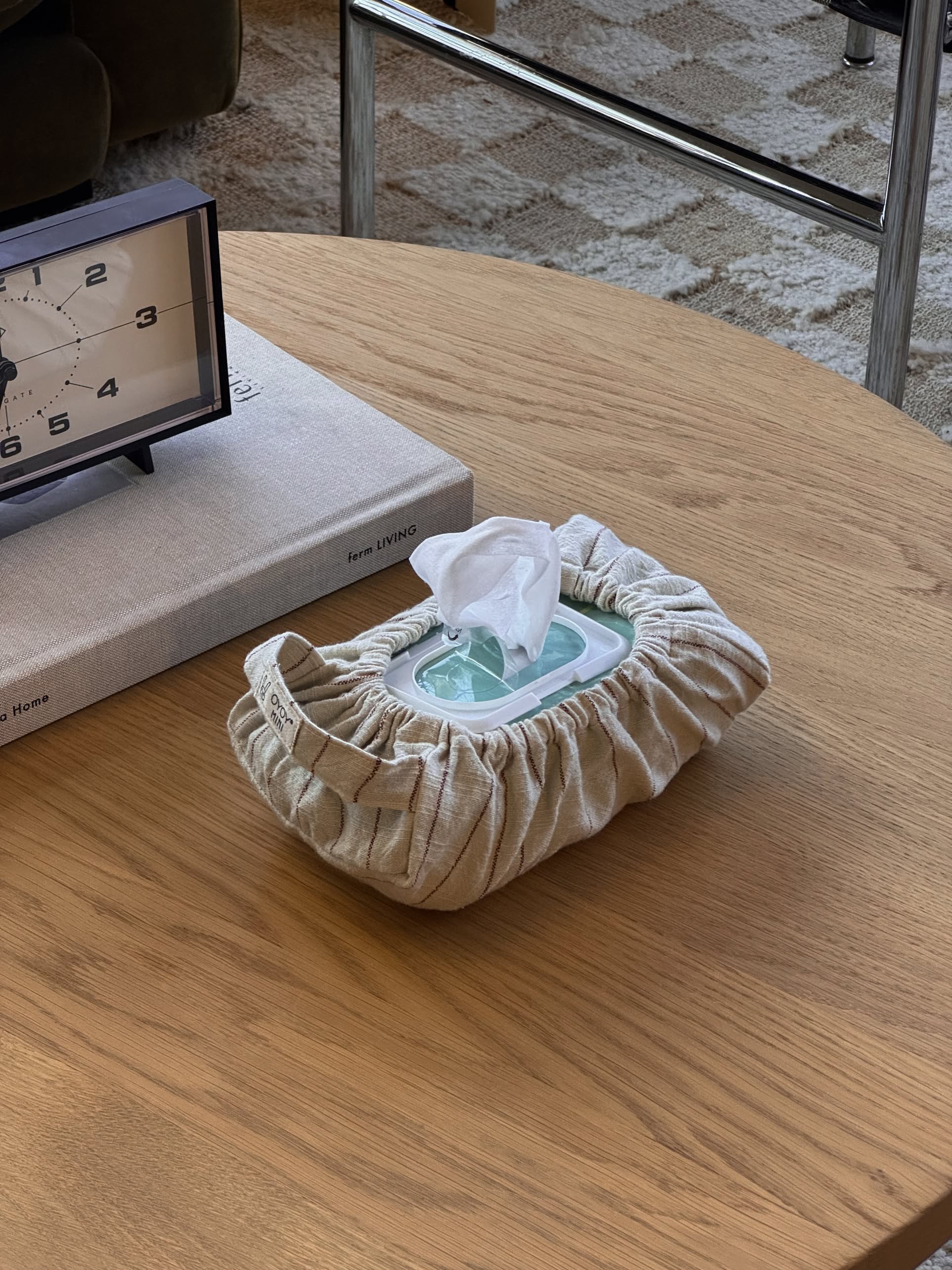The OYOY Living Design Wet Wipes Cover in Nutmeg sits on a wooden table beside a book and clock, with a wipe ready for use, giving the cozy, modern interior a stylish touch.