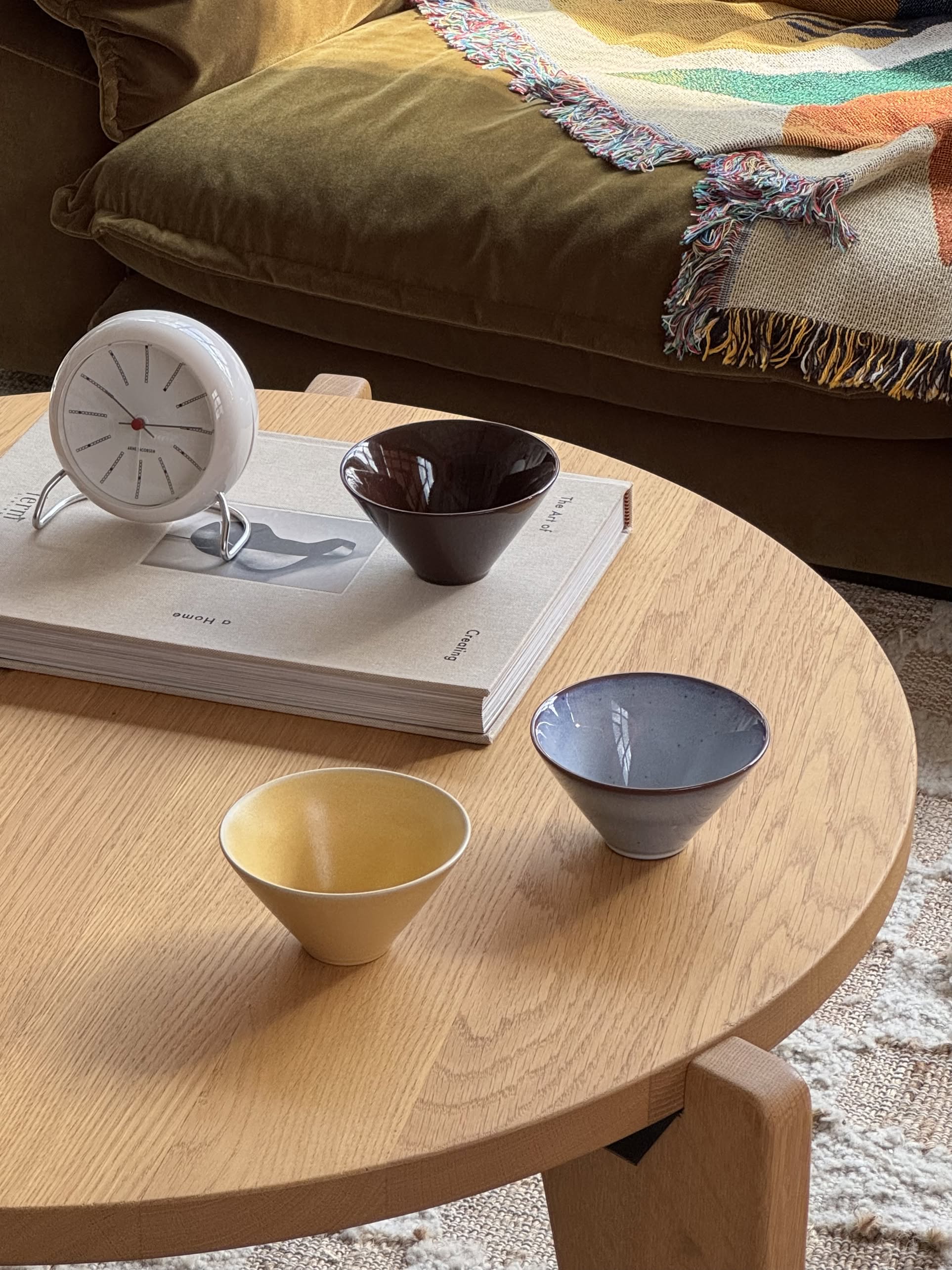 A round wooden table holds the OYOY Living Design Yuka Bowl, Set of 3 - Dark Terracotta, plus a clock and book. Behind the table, a colorful fringed blanket adds decorative flair in front of a couch.