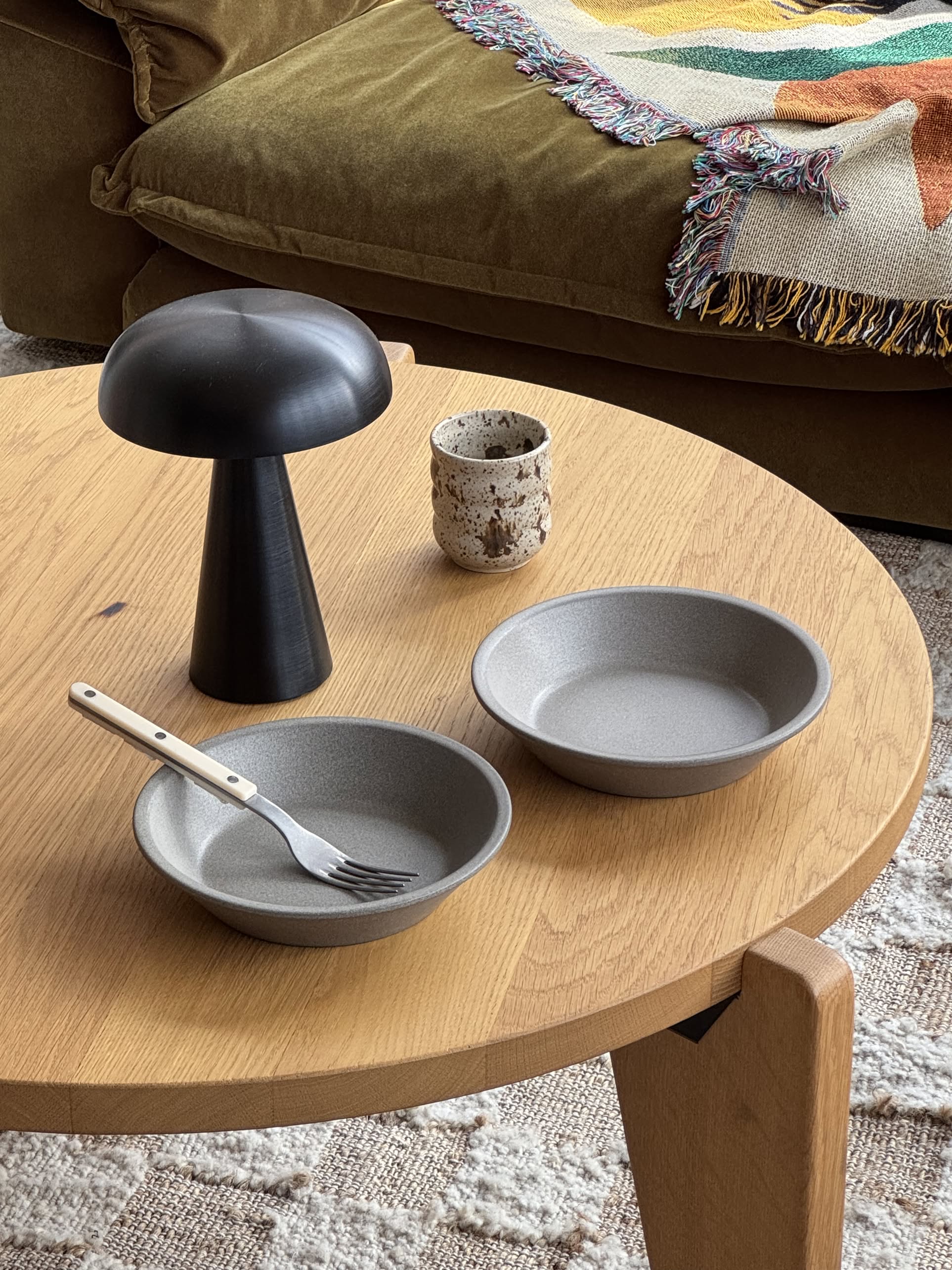 A round wooden table set with OYOY Living Design Yuka Deep Plates (Set of 2, Stone), a fork, rustic cup, and black mushroom lamp. In the background, a green sofa features a fringed patterned throw blanket.