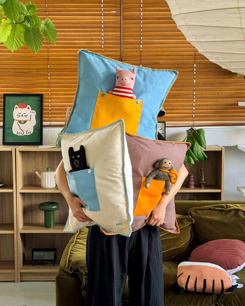 A person holds three Ordinary Daily Design Pocket Cushion Covers in brown and orange, each featuring a small pocket containing a plush stuffed animal, in a cozy, sunlit living room with wooden blinds and playful decor.