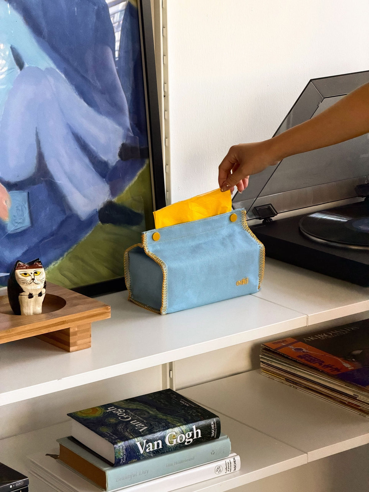 A hand places a yellow envelope into a blue fabric organizer on a white shelf, where books, magazines, a cat figurine, the Ordinary Daily Design Tissue Case - Blue/Yellow Stitch by Ordinary Daily Design, and part of a painting are visible nearby.