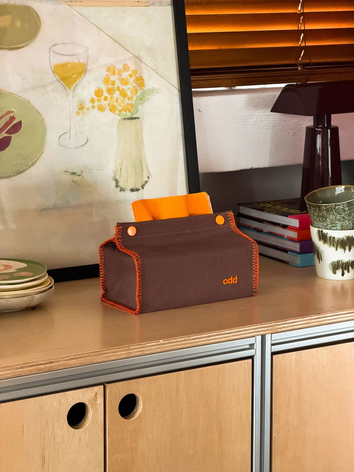An Ordinary Daily Design Tissue Case in brown with orange stitching, labeled “odd,” rests on a wooden cabinet beside stacked books, a cup, and dishes, with orange blinds and artwork in the background.