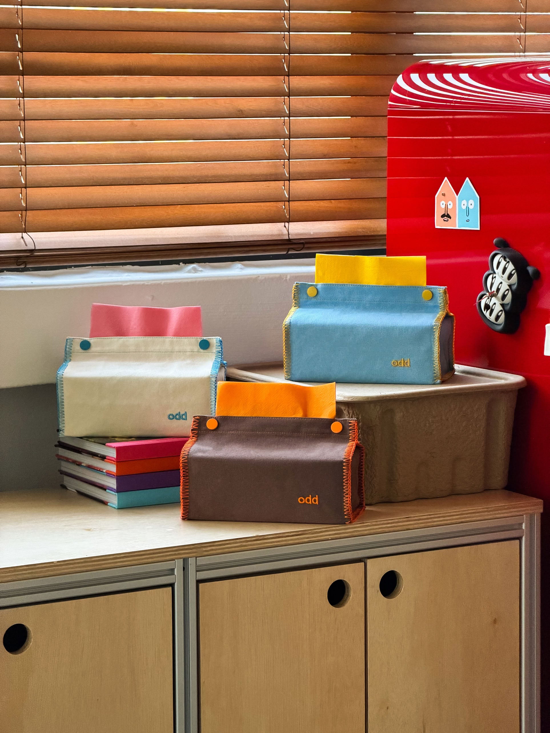 Four Ordinary Daily Design Tissue Cases in brown with orange stitching, featuring button closures, are displayed on a wooden cabinet next to a stack of books and a red refrigerator, in a room with wooden blinds.