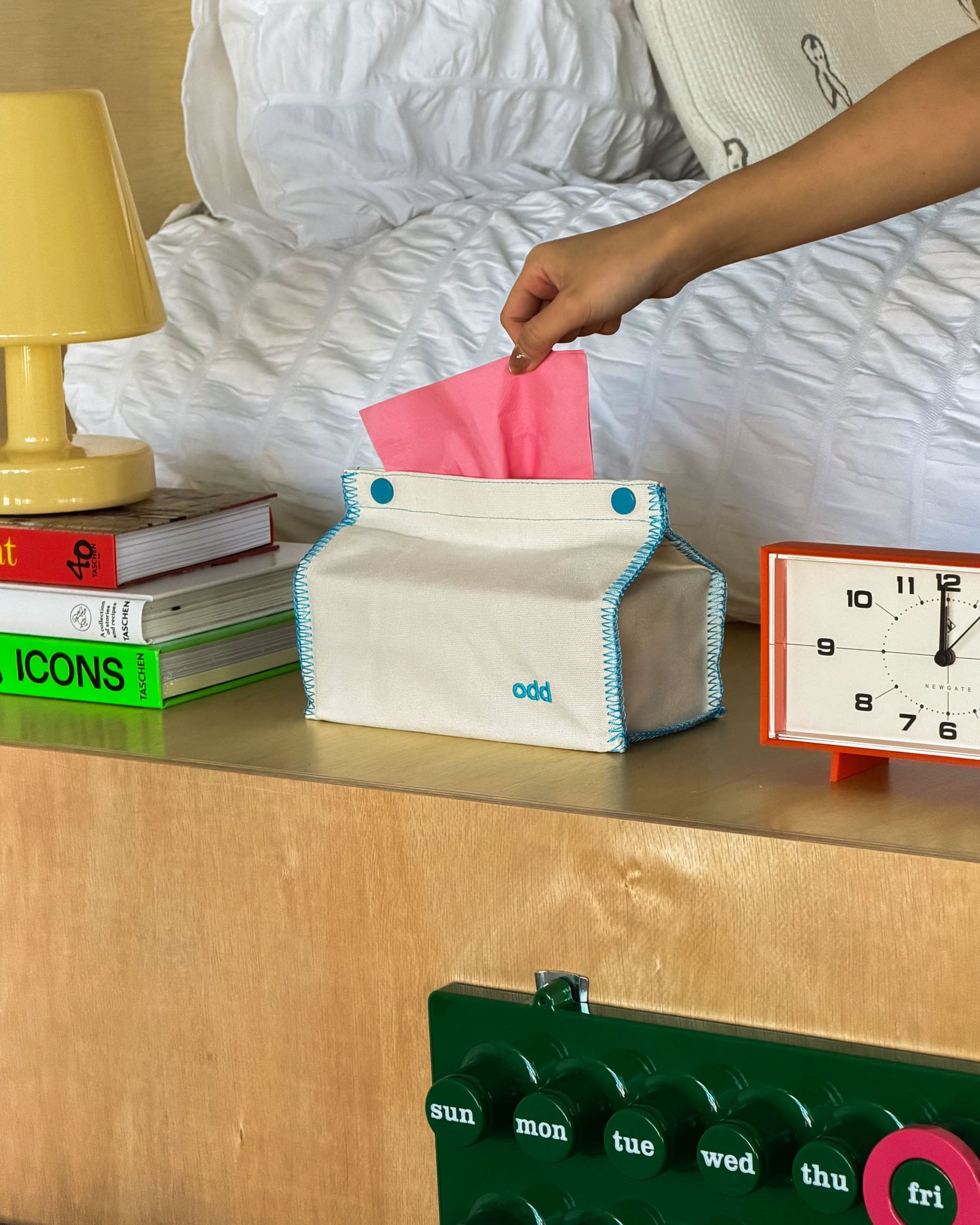 A hand pulls a tissue from the Ordinary Daily Design Tissue Case in Cream/Blue Stitch by Ordinary Daily Design, which sits on a bedside table next to books, a yellow lamp, and an orange clock showing 8:20.