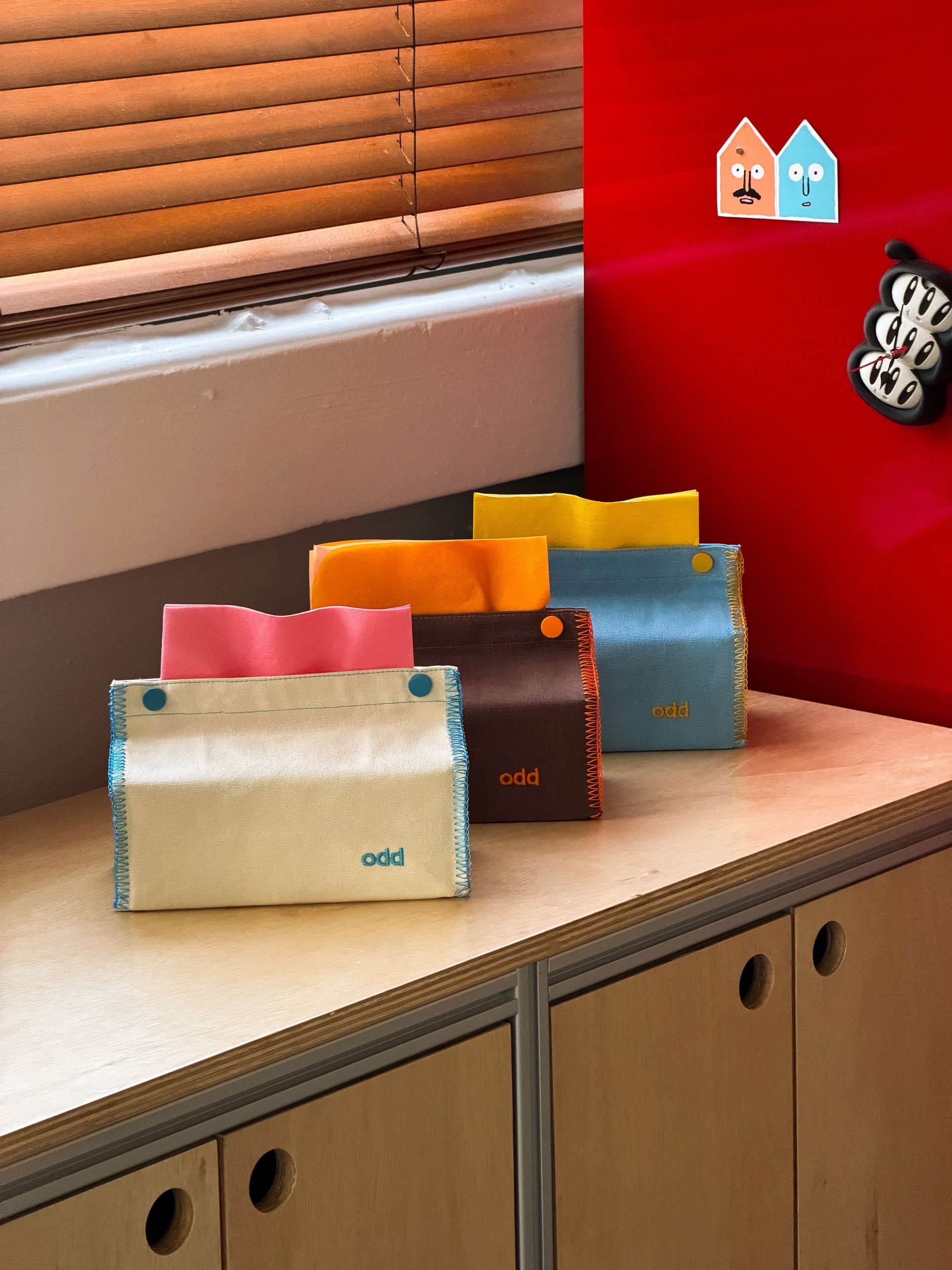 Four colorful wallets, each with a button closure, are arranged on a wooden cabinet next to the Ordinary Daily Design Tissue Case in Cream/Blue Stitch by Ordinary Daily Design. A bright red wall and two cartoon house stickers can be seen in the background near a window with wooden blinds.