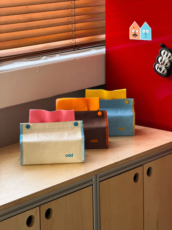 Four colorful wallets, each with a button closure, are arranged on a wooden cabinet next to the Ordinary Daily Design Tissue Case in Cream/Blue Stitch by Ordinary Daily Design. A bright red wall and two cartoon house stickers can be seen in the background near a window with wooden blinds.
