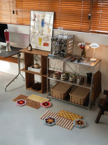 A cozy corner features a coffee machine, cups, lamp, decorative items, and an art print displayed on shelves; two woven baskets sit underneath; the Ordinary Daily Design Deconstructed: Flower Rug from Ordinary Daily Design adds a vibrant touch to the floor; blinds cover the window behind.