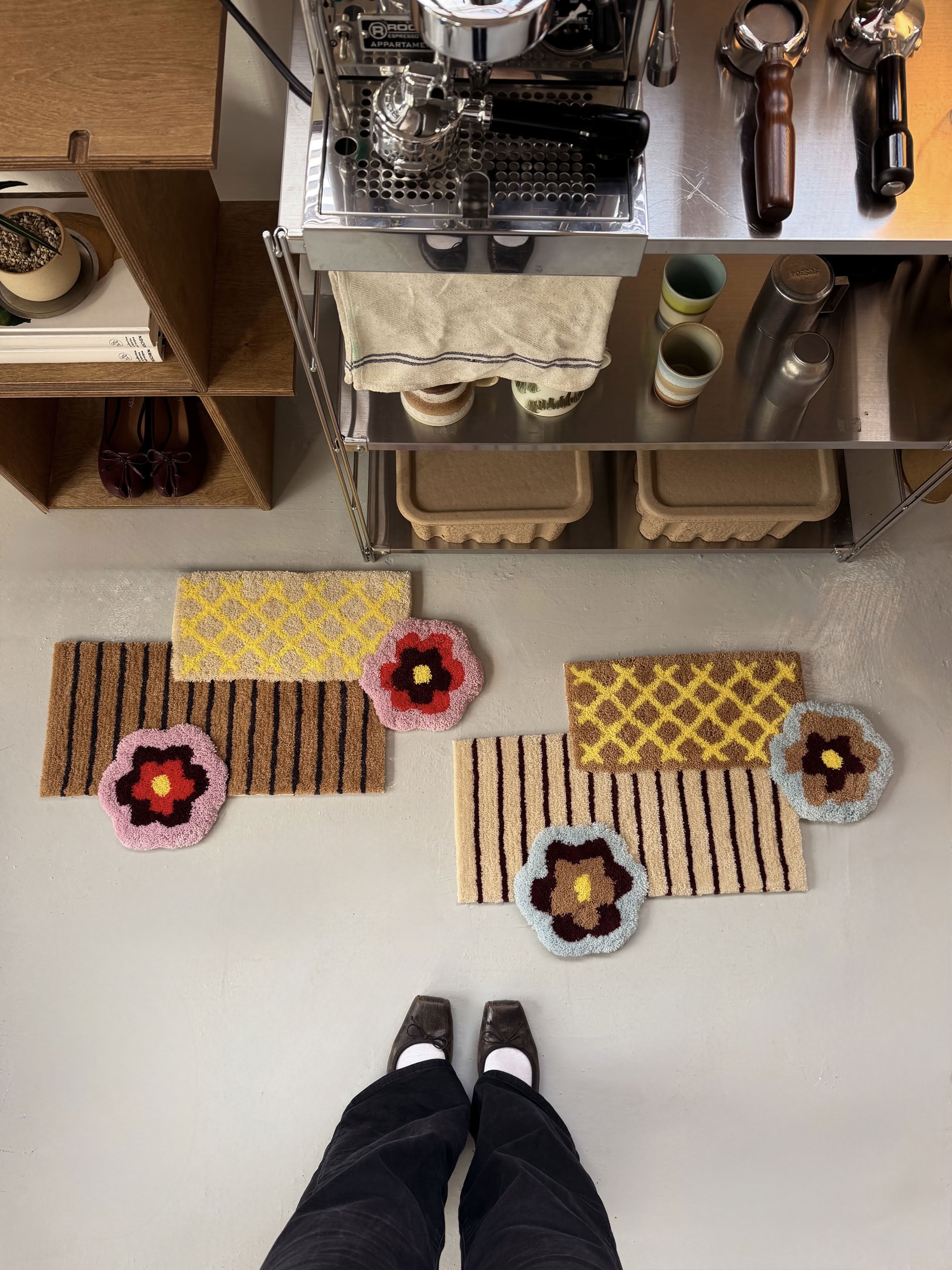 A person stands on a light floor in front of several colorful doormats, including the Ordinary Daily Design Deconstructed: Flower Rug by Ordinary Daily Design—a pink rug with floral patterns—alongside other geometric designs near a coffee station outfitted with cups, dishes, and a coffee machine. Black shoes and dark pants are visible at the bottom of the image.