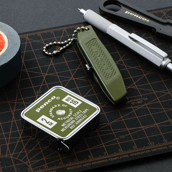 The Penco Pocket Measure, a compact green and white tape measure from Penco, rests on a black grid cutting mat alongside a utility knife, pen, and tape roll. All tools are neatly arranged for an organized workspace.