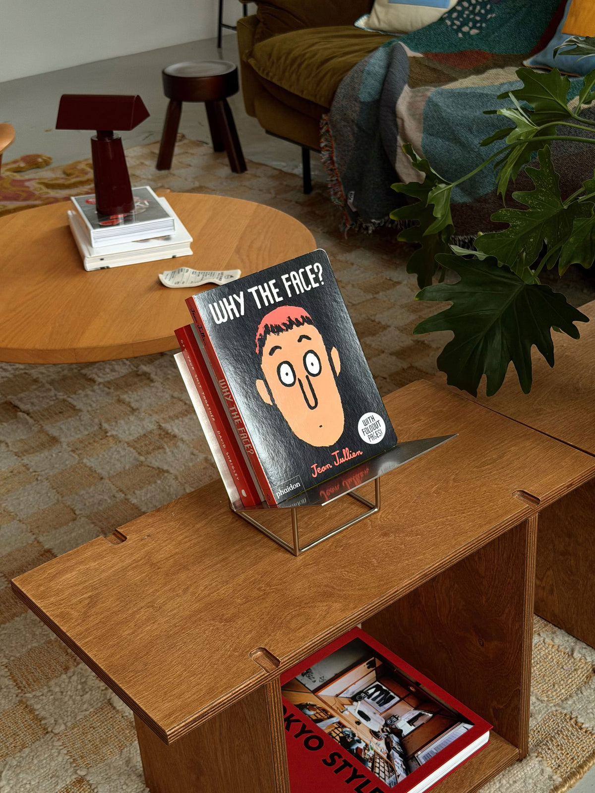 A cozy room with a wooden table and bookshelf features Phaidon’s Why The Face?, a board book by Phaidon designed to help children develop emotional awareness through playful exploration of facial expressions. A red lamp, stacks of books, and a blanket draped over the sofa add warmth to the inviting scene.