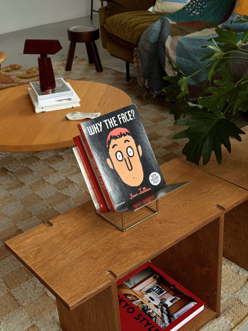 A cozy room with a wooden table and bookshelf features Phaidon’s Why The Face?, a board book by Phaidon designed to help children develop emotional awareness through playful exploration of facial expressions. A red lamp, stacks of books, and a blanket draped over the sofa add warmth to the inviting scene.