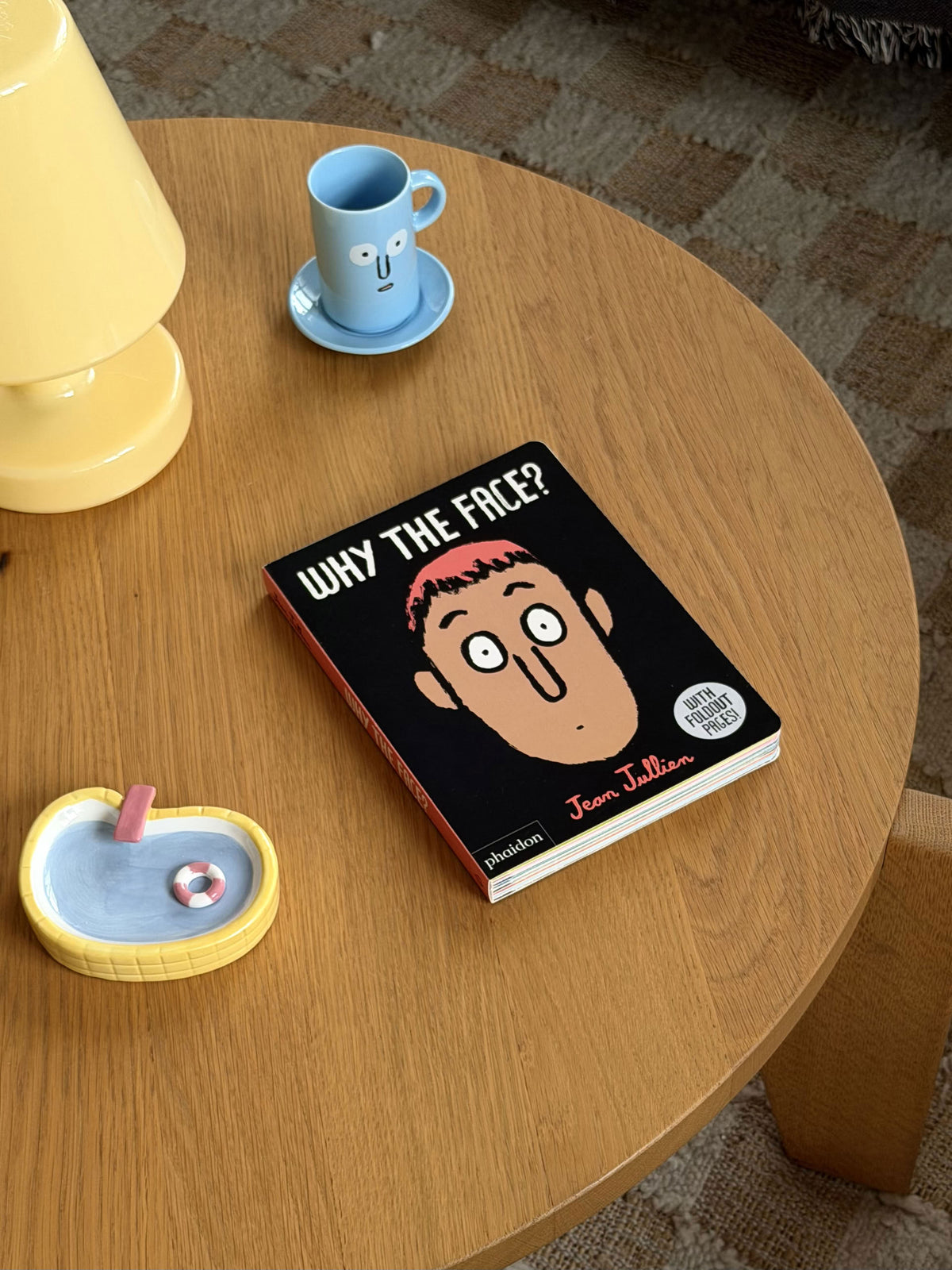A round wooden table with a yellow lamp, a small blue cup and saucer featuring a face showing different expressions, a yellow ashtray with a cigarette and lighter, and Phaidon Why The Face? by Phaidon—a board book that playfully encourages emotional awareness.