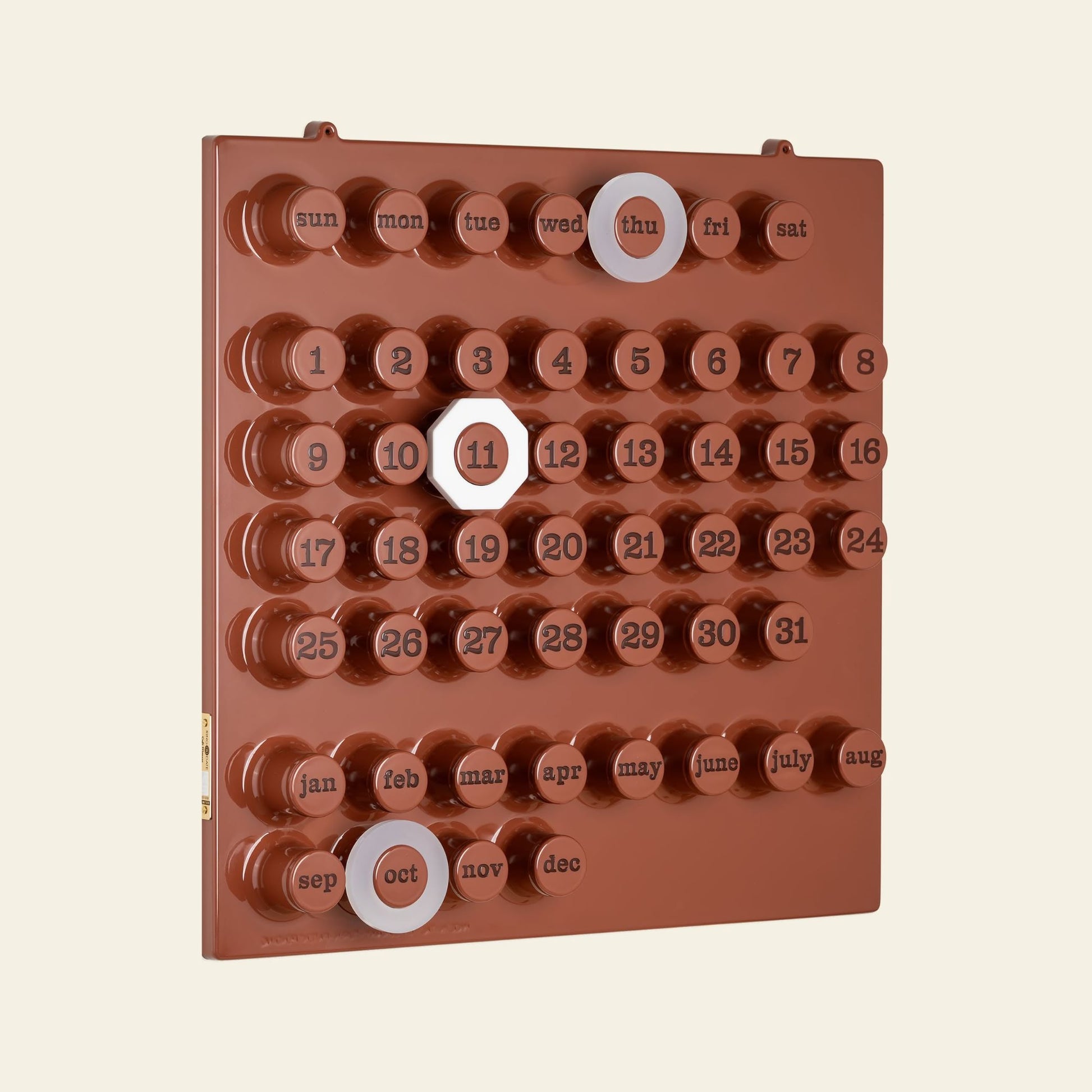 The Ring-A-Date Limited Edition Wall Calendar - Cafè Mousse features a light beige background and brown design with round buttons for days, dates, and months. White markers highlight Thursday, the 11th, and October.