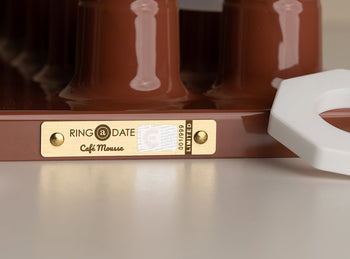 A close-up of the Ring-A-Date Limited Edition Wall Calendar in Café Mousse, featuring a gold and white tag, two screws, and a partially visible white handle on a light surface.