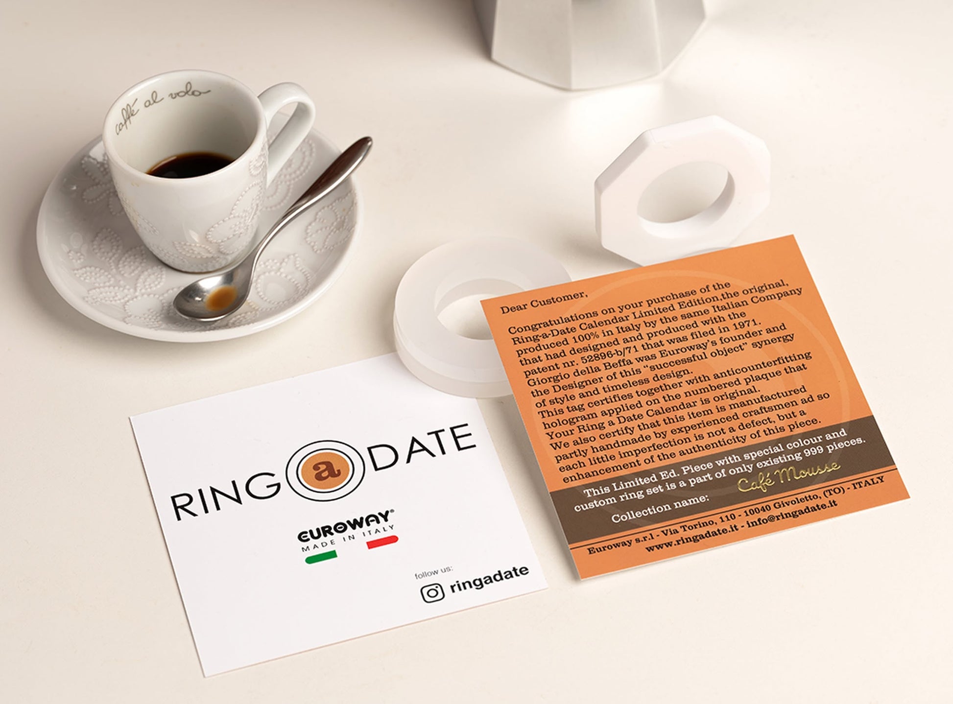 A cup of coffee on a saucer with a spoon sits beside a hexagonal white ring, RINGADATE logo card, orange info card, and the Ring-A-Date Limited Edition Wall Calendar - Cafè Mousse by Ring-A-Date on a white surface.