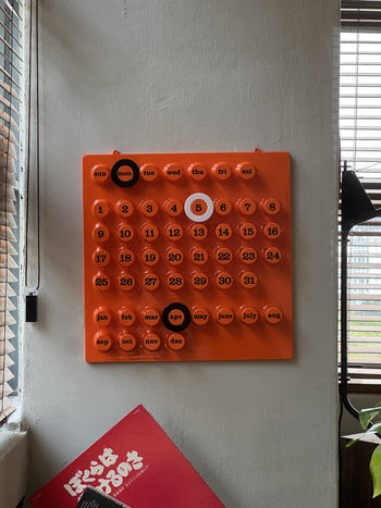 The Ring-A-Date Wall Calendar by Ring-A-Date is an orange retro calendar with removable circular markers. Today’s date, Monday April 5, is highlighted with black and white markers. This perpetual calendar hangs on a white wall.
