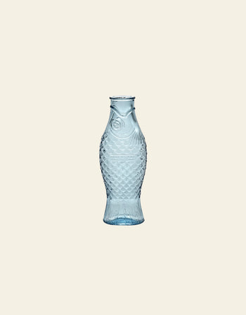 The Serax Fish & Fish Carafe by Serax, featuring textured scales and fins in transparent light blue glass, stands out as an eye-catching tableware piece against a plain beige background.
