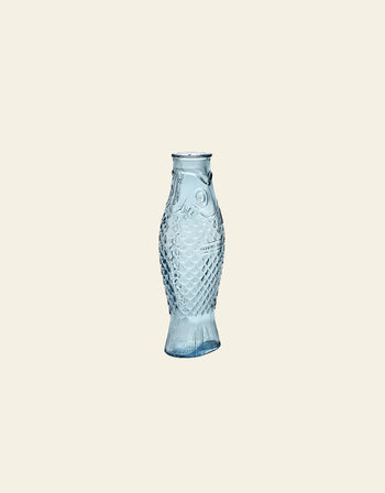 The Serax Fish & Fish Carafe by Serax, with its blue-tinted glass, detailed scales, and fins, stands out as a unique piece of tableware set against a plain light beige background.