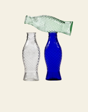 The Serax Fish & Fish Carafe set by Serax features three glass carafes—clear, blue, and light green—shaped like fish. The clear and blue stand upright, while the green rests horizontally on top for a unique table display.