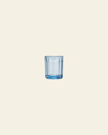 A blue-tinted glass tumbler, half-filled with water, is centered on a plain light beige background next to a green Serax Fish & Fish Glass M carafe by Serax.