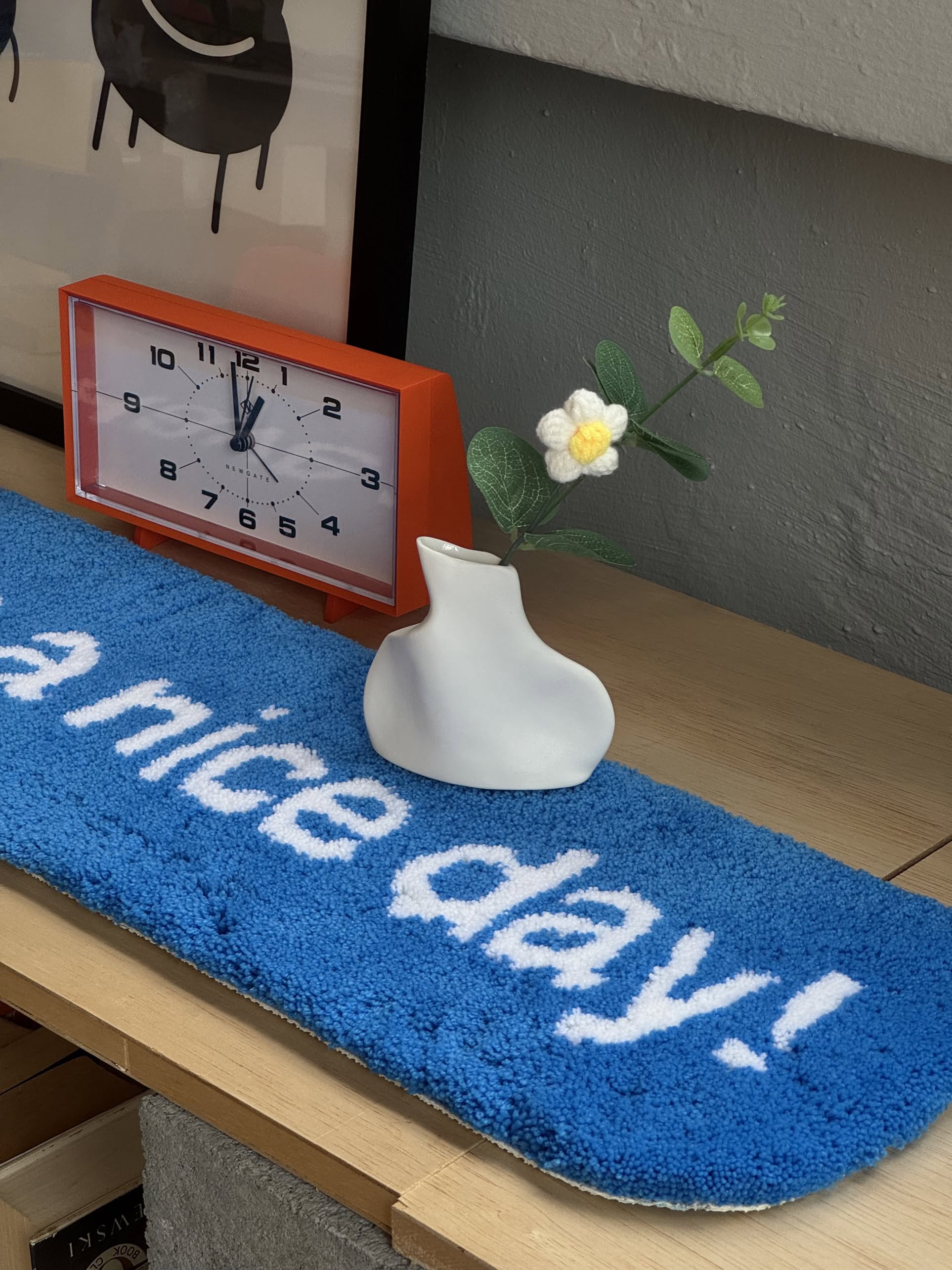 A red-framed clock showing 11:59 sits on wood beside a Serax Perfect Imperfection Flower Vase N°5 in white, holding a small flower. In front, a blue mat displays a nice day! in white letters.