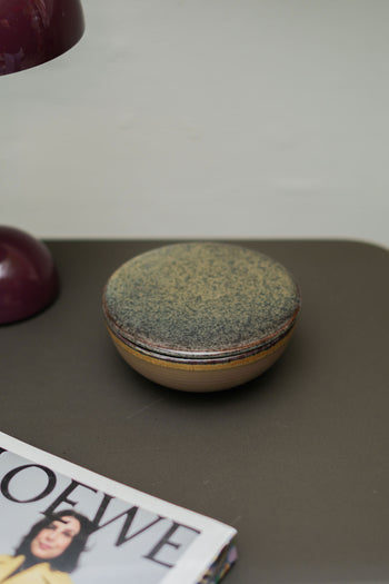 The Serax Surface Bowl & Lid - Indi Grey by Serax, a round, handcrafted piece, sits on a dark surface beside a burgundy object and the edge of a magazine.