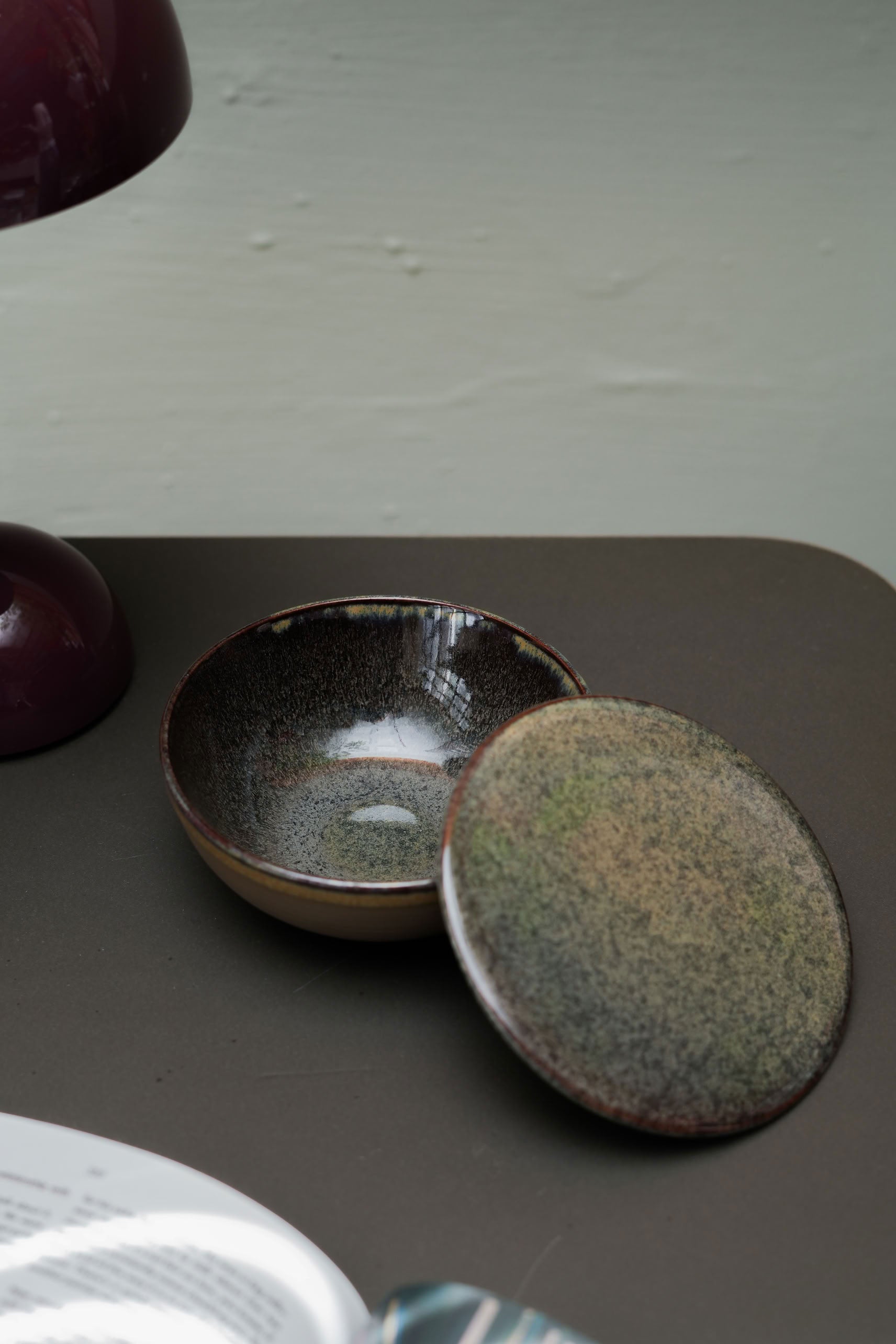 The Serax Surface Bowl & Lid in Indi Grey by Serax sits on a brown surface with its matching lid slightly open. A portion of a purple lamp and the edge of a book are also visible nearby.