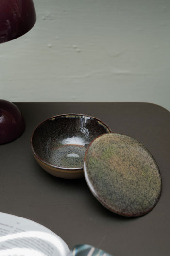 The Serax Surface Bowl & Lid in Indi Grey by Serax sits on a brown surface with its matching lid slightly open. A portion of a purple lamp and the edge of a book are also visible nearby.