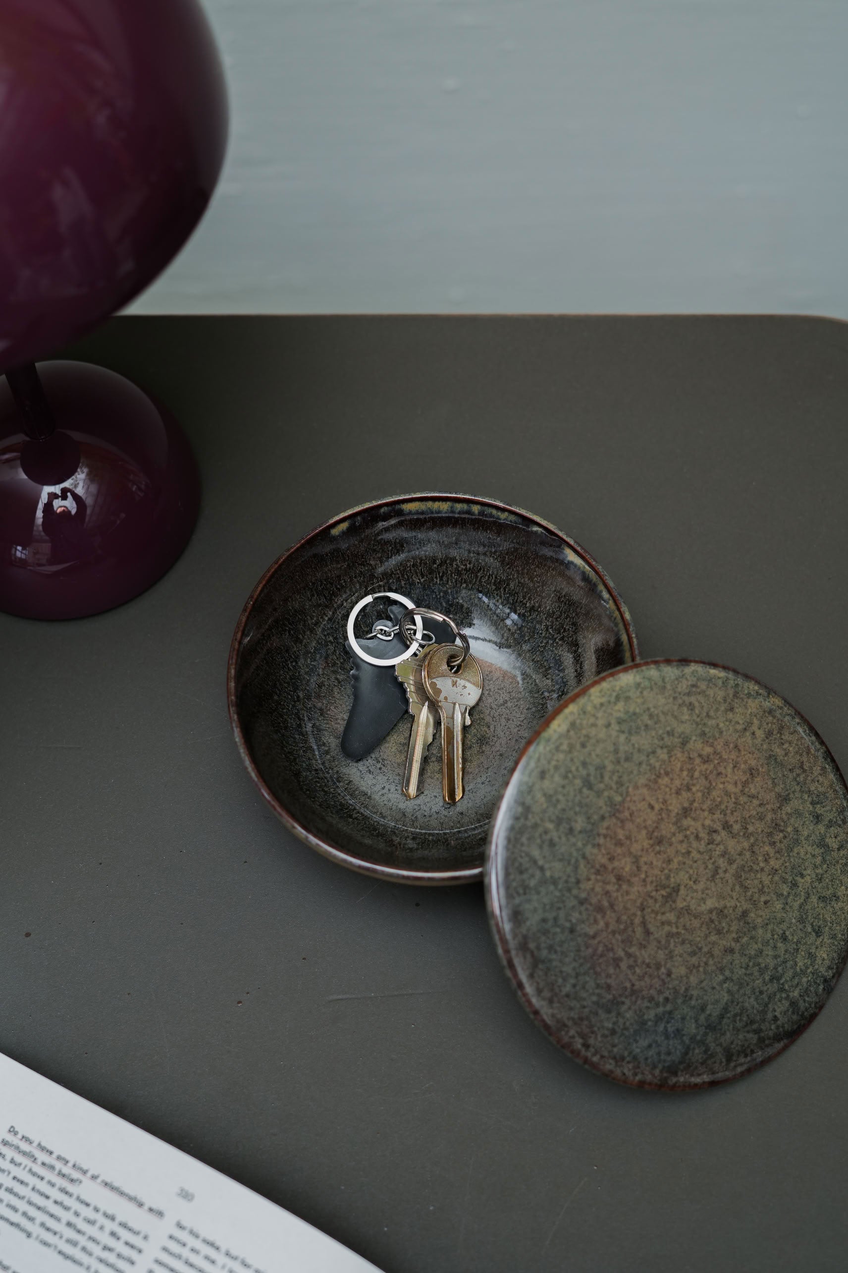 The Serax Surface Bowl & Lid - Indi Grey by Serax holds keys with a black keychain, arranged on a dark surface beside a purple lamp and an open book or magazine.