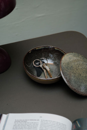 The Serax Surface Bowl & Lid - Indi Grey by Serax holds several keys and a black key fob, resting on a dark surface next to an open book.