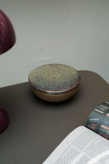 The Serax Surface Bowl & Lid in Indi Grey, a round handcrafted ceramic piece, rests on a dark surface beside a purple lamp and an open magazine.