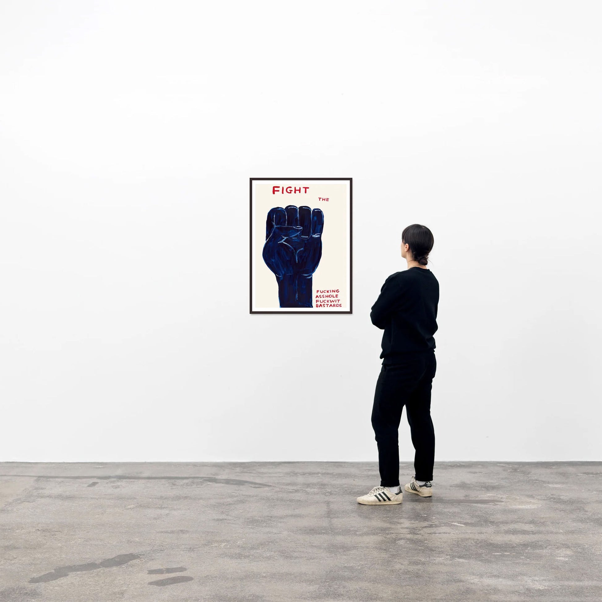 In a minimalist gallery, a person admires the Shrig Shop David Shrigley - Fight (50x70) poster, featuring a large dark blue fist with the word FIGHT in bold red letters at the top.