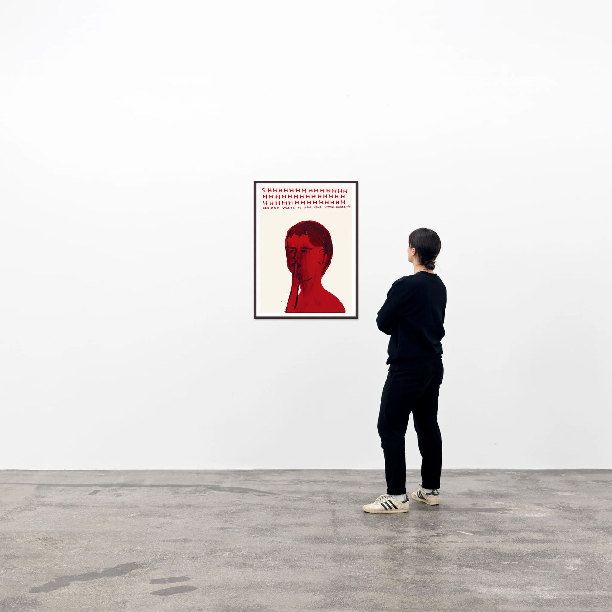 A person in black stands in a gallery, admiring the Shrig Shop David Shrigley - Shhh (50x70) poster: a red-toned portrait with text above the head, displayed on a white wall above the gray concrete floor.