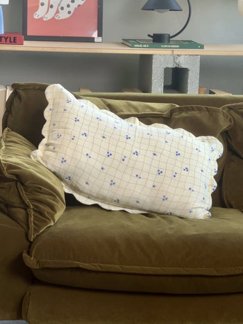 A green velvet couch with Soft Spot Soft Petal Cushion Covers Rectangle by Soft Spot. The white cushion covers feature a blue floral grid pattern. Behind the couch are shelves with books, a lamp, and colorful artwork.