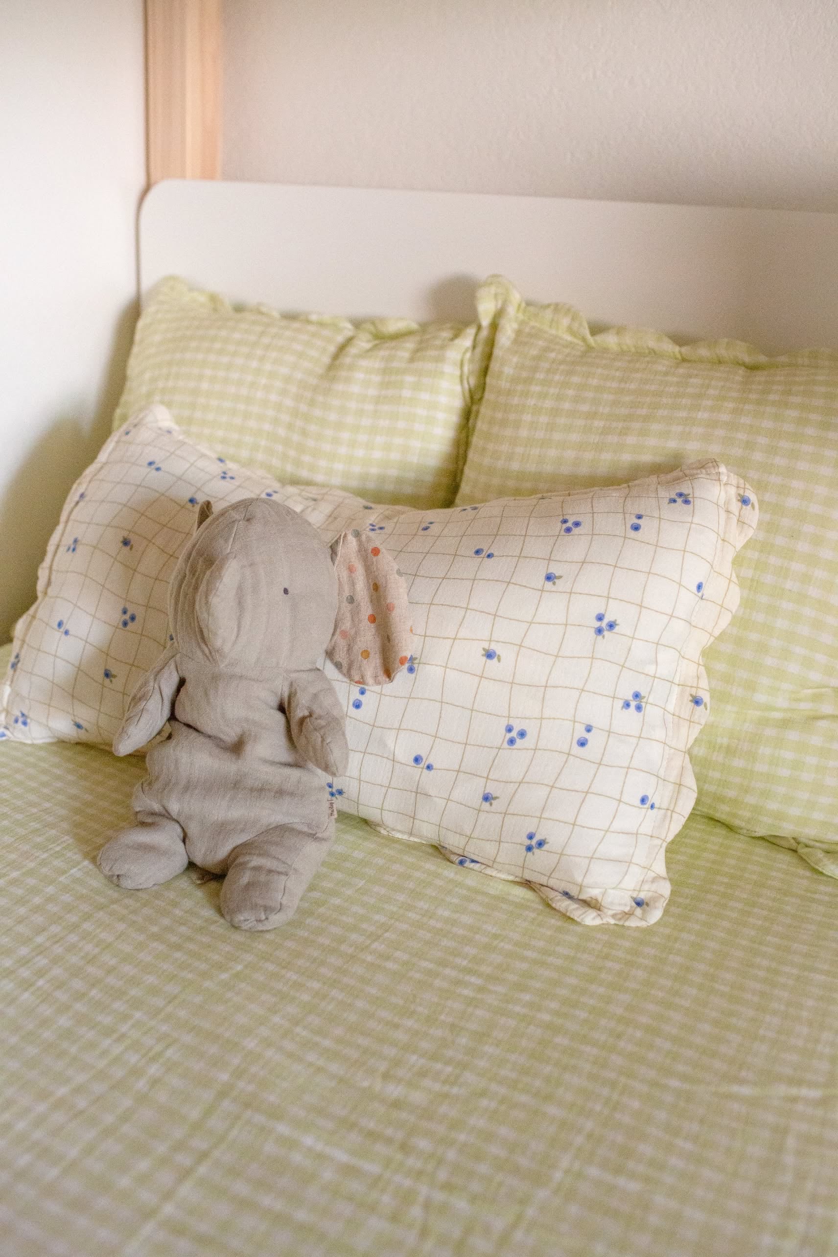 A stuffed bunny toy sits on a neatly made bed with light green checkered sheets and two Soft Spot Soft Petal Cushion Covers Rectangle by Soft Spot—one in green and the other with a cream and blue floral grid design.