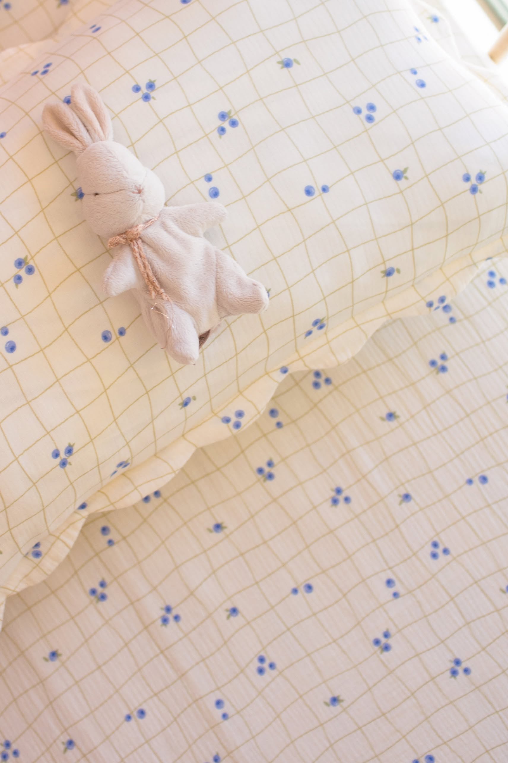 A small, plush bunny toy rests on a Soft Spot Soft Petal Rectangle Cushion Cover featuring a yellow grid and blue flowers, matching the bedding underneath.